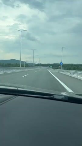 A first-person point-of-view driving video shows a car traveling down a multi-lane highway on a cloudy day. The sky is filled with grey clouds, and the road ahead is empty of other vehicles. Streetlights line the highway, and green road signs indicating directions are visible in the distance. Lush green trees and vegetation flank both sides of the road, and mountains are visible in the far background. The car's dashboard and windshield are visible in the foreground.