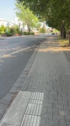 A red car drives away from the camera on a road with a sidewalk on the right. The sidewalk is made of gray pavers and has a tactile paving section. The road is asphalt and has white lane markings. Trees with green leaves line the right side of the road, casting shadows on the sidewalk. Buildings and more trees are visible on the left side of the road in the background. The sky is blue and clear.