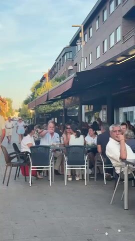 The video shows an outdoor seating area of a restaurant or cafe. Several people are seated at tables, some are talking and eating. In the background, people are walking along a sidewalk. The setting appears to be a city street with buildings lining one side of the street and trees and foliage. The lighting suggests it is late afternoon or early evening. The weather appears to be pleasant. There is a baby in a stroller in the foreground. One man is talking on his phone. The overall atmosphere is relaxed and social.