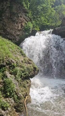 A waterfall cascades down a rocky cliff face. Lush green vegetation, including trees and moss, covers the surrounding rocks. The water is white and frothy as it plunges into a pool below. Sunlight filters through the trees, casting dappled shadows on the scene. The sky is blue with scattered white clouds.