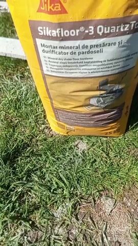 Here is a concise description of the video:

The video shows a bag of SikaFloor-3 QuartzTop mineral dry shake floor hardener sitting on a grassy area. In the background, a person is using a power trowel to smooth concrete. The camera focuses on the Sika product packaging. The environment appears to be a construction site or area where concrete work is being done.