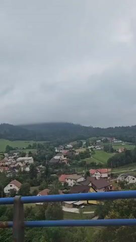 The video shows a scenic view of a village nestled in a valley with rolling hills covered in trees and vegetation. The sky is overcast with clouds. In the foreground, there is a blue railing, suggesting the viewpoint is from a bridge or elevated platform. The village consists of numerous houses with pitched roofs, varying in color. Green fields and trees are interspersed among the buildings. The overall atmosphere is calm and serene.