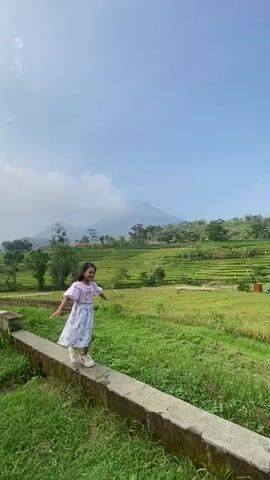 The video shows a young girl walking on a low concrete wall in a rural, scenic environment. She wears a light purple and floral patterned dress with short, puffy sleeves and light pink shoes. She has short, dark hair and is smiling. The girl walks carefully, using her arms for balance, against the backdrop of vibrant green rice terraces that stretch up to the base of a mountain, which is partially obscured by clouds. The sky is light blue with scattered white clouds. The concrete wall is covered with green grass and some weeds. In the background, there are a few small structures among the rice terraces.