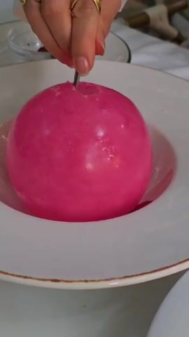 Here is a description of the video:

A person uses a spoon to puncture a large, pink, spherical dessert sitting in a white bowl. The shell cracks, revealing a scoop of white ice cream drizzled with caramel sauce and garnished with rose petals. The person then uses the spoon to place the pink shell fragments back on top of the ice cream. The scene is well-lit.