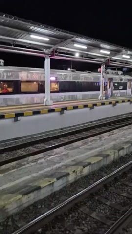 A train is parked at a station at night. The train is silver with pink accents. It is illuminated by overhead lights. The station platform is visible in the foreground, with train tracks running alongside it. The sky is dark and clear. There are no people visible in the video.