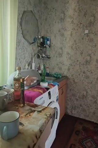 The video pans across a small room that appears to be a kitchen or utility area. On the left, a washing machine is visible next to a countertop with a sink. Various items, including bottles, a sponge, and what looks like a pink blanket, are placed on the counter and washing machine. A bottle with a label is on a cluttered surface in the foreground. The walls are covered in a grey, textured wallpaper. To the right, a bright teal door is prominent, with a patterned rug on the floor in front of it. The rug features geometric designs in red, black, and white. Towards the right side of the frame, coats and bags are hanging on a wall. The lighting is dim, suggesting an indoor environment.