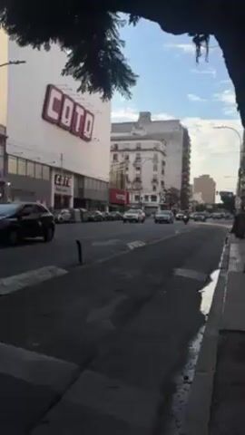 The video shows a city street with cars driving on the road. On the left side of the road is a large building with the word "COTO" in red letters on the side. There are other buildings visible in the background. A motorcycle rides on the street. The sky is blue with some clouds. A tree branch hangs into the frame in the upper part of the video. There are streetlights and a sidewalk on the right side of the road.