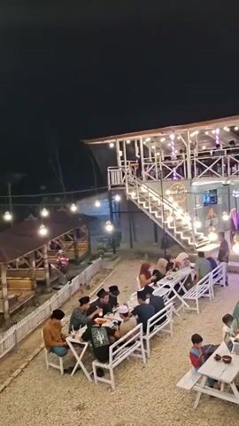 The video shows an outdoor dining area at night, illuminated by string lights and individual light bulbs. A two-story building, likely a restaurant or cafe, is visible with people dining on both levels. Stairs lead up to the second level. In the foreground, several groups of people are seated at white picnic tables and benches, some eating and others conversing. There are also motorcycles parked in the background near the building. The ground is covered with gravel. The overall atmosphere appears to be casual and social.