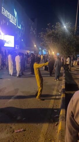 At night, a street scene unfolds in front of a brightly lit building displaying the text "EMPORIUM Mall by Nishat Group". Numerous people, primarily men dressed in traditional South Asian attire like shalwar kameez, are gathered on the street and sidewalk. The ground is dark asphalt, and a yellow-painted curb runs along the right side of the frame, showing some litter. Streetlights cast long shadows and illuminate the area. In the foreground, a man in a yellow shalwar kameez stands with his back to the camera, extending his right arm and pointing forward. Other individuals are seen walking, standing, and conversing in small groups. The overall atmosphere suggests a busy evening outside a shopping center.