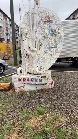 The video shows a sculpture in an outdoor urban area, likely a park or street side. The sculpture is painted white and heavily splattered with various colors of paint, giving it an artistic and slightly vandalized appearance. It depicts an artist standing next to a large, circular palette with brushes pointing upward. The base of the sculpture has the word "КРАСКИ" (which translates to "Paints") written on it in red paint, which also appears dripped and smeared. The surrounding environment includes a playground with yellow and red structures, a parked silver car, a white box truck, a building with visible windows, and a patch of gravelly ground. The sky appears overcast, suggesting a cloudy day. There are no people clearly visible in the video.