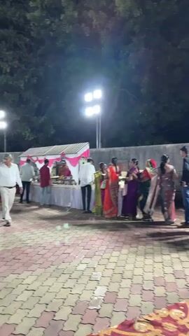 The video shows a gathering of people outdoors at night, possibly at an event. Several individuals, including women wearing saris and men in shirts and pants, stand in a line near a table covered with food and decorations. A few men are walking and chatting with each other. Overhead lighting illuminates the scene, casting shadows on the brick-patterned ground. In the background, there's a tree and what appears to be a wall. A carpet or rug is partially visible on the ground in the foreground.