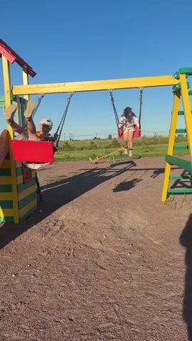 Two children are swinging on a playground. The playground structure is yellow and green with a red roof. The ground is covered in gravel. The sky is clear and blue. The child on the left is wearing a blue shirt, orange shorts, and a white baseball cap. The child on the right is wearing a white shirt, patterned shorts, and a pink headband. They are both swinging back and forth. The sun is casting shadows on the ground.