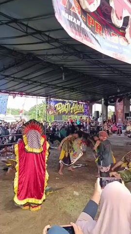 A group of people are gathered under a large tent to watch a performance. In the foreground, two people are holding up phones to record. The performance features several individuals dressed in colorful costumes. One performer wears a large, red, hooded costume with yellow trim. Several other performers are dressed in green and yellow outfits. Some of the performers are dancing or moving in a way that suggests a ritual or traditional dance. There are banners hanging in the background, one of which reads "PANJI ANOM". The ground is dirt. There is an audience seated in rows behind the performers.
