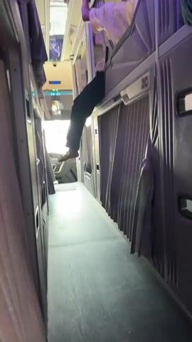 The video captures the interior of a sleeper bus. The camera moves down a narrow aisle between rows of sleeping berths. On the left, the berths are visible with gray curtains. On the right, the berths are also visible with similar gray curtains and white trim. A person's legs and feet are seen hanging out of an upper berth on the left side of the aisle, with their feet dangling towards the aisle. The person is wearing dark pants and appears to be in the process of getting in or out of the berth. The aisle floor is dark and appears to be made of wood or a similar material. The lighting is dim, with some purple and white lights visible from above and within the berths. Further down the aisle, the driver's cabin is partially visible through a window. The video pans to the right, showing more berths with closed curtains. One berth on the right has its curtain slightly open, revealing a bed with a turquoise comforter and a small bedside table. The overall impression is of a cramped but functional sleeping arrangement on a long-distance bus.