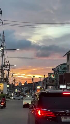 The video captures a street scene at dusk, with a vibrant sunset visible in the distance. The sky is filled with dramatic clouds tinged with orange, pink, and purple hues, illuminated by the setting sun. A black minivan is in the foreground, driving on a road lined with buildings and streetlights. There are other vehicles and motorcycles on the road, suggesting moderate traffic. Telephone poles with a complex network of wires crisscross the sky, adding to the urban atmosphere. Signs for businesses, including a 7-Eleven, are visible on the buildings. The overall impression is one of a bustling street as day transitions into night, bathed in the warm glow of a sunset.