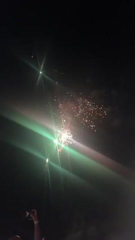 The video shows a nighttime fireworks display. A hand holding a phone is visible in the lower left corner of the screen, capturing the event. Fireworks explode in bursts of red and white light against the dark sky, with some green sparks also present. Smoke trails from the explosions, and bright beams of light from the fireworks cut through the darkness.