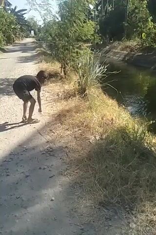 A young boy is standing on a dirt path next to a canal. He is wearing a black t-shirt and shorts. He is holding a small object in his right hand, and he is in the process of throwing it into the canal. The canal is filled with water and surrounded by tall grass and trees. The sun is shining, casting shadows on the path. The boy is looking towards the canal as he throws the object.