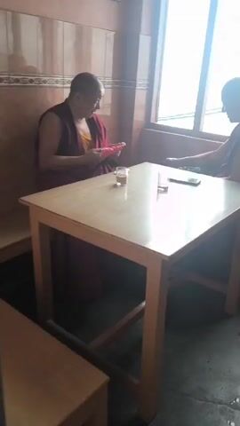 The video opens with a close-up of two individuals dressed in maroon robes sitting at a wooden table. One person on the left is holding a pink object, possibly knitting. There are two glasses with liquid, likely tea, and a smartphone on the table. The scene is indoors, near a window. The camera then pans to the right, showing the second person with their hands clasped, looking out the window. The view through the window reveals a misty, possibly rainy, outdoor environment. There is a black umbrella leaning against the wall. The scene transitions to a wider view of the outdoors, showing a street with several auto-rickshaws parked. Buildings with green roofs are visible, along with some trees and electrical wires. A yellow billboard with an image of a woman is also present. The overall atmosphere suggests a cloudy or rainy day in a town.