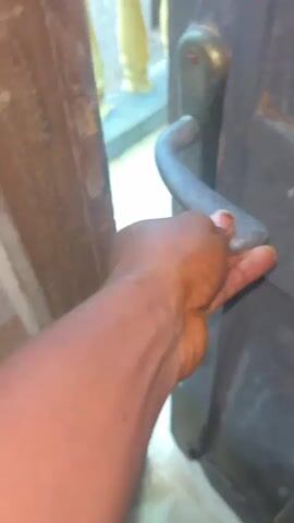 The media file shows a first-person perspective of someone approaching and opening a wooden door. The scene appears to be outdoors, with a tiled floor visible in the foreground and a concrete railing with decorative yellow pillars in the background. A yellow plastic container, possibly a jerrycan, sits on the railing. Beyond the railing, some construction or building materials are visible, suggesting an unfinished or developing area. The ground outside the railing appears wet and muddy, hinting at recent rain or damp conditions.

The subject is an individual's arm and hand as they reach for and grasp a metal door handle. The hand is dark-skinned, and the arm shows prominent veins. The door itself is dark and appears weathered, with a tarnished brass lock mechanism. The action of opening the door suggests the person is entering or exiting a building.

The lighting suggests it is daytime, but the quality of light is not distinct enough to pinpoint the exact time. There are no clear indicators of the specific weather, other than the dampness on the ground. The location context of Ikorodu, Nigeria, aligns with the visual cues of the outdoor environment, the type of building materials, and the general aesthetic of the scene. No text is clearly visible in the media file. The overall tone is one of simple, everyday action.