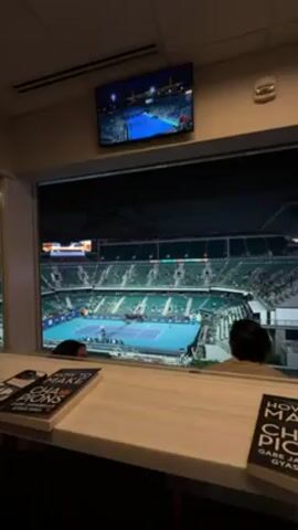 The video shows an indoor view from a room overlooking a tennis court in a large stadium. The camera pans down from a television on the wall playing a game to reveal the stadium through a large window. People can be seen in the stadium stands watching the tennis game. Two people are partially visible from inside the room in front of the window. Two copies of a book titled "How to Make Champions" are on the table in front of the window.