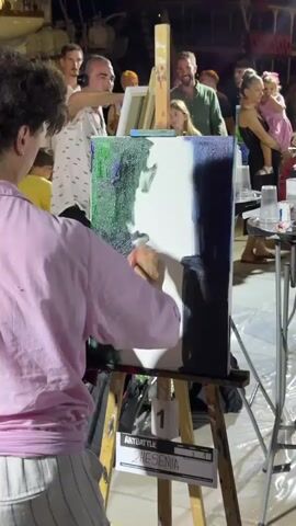 A person in a pink shirt is painting on a canvas placed on an easel. The person is using a paintbrush to add strokes of dark blue paint to the right side of the canvas. The canvas also has green and white paint on it. In the background, there are several people watching. Some of them are adults, and one is a child being held by an adult. The scene appears to be outdoors at night, possibly at an art event or competition, given the presence of easels, paints, and a sign that says "ART BATTLE" and "THESENIA".
