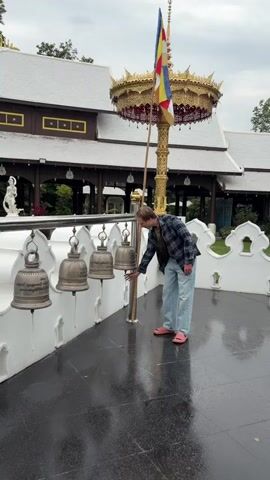 A person wearing a plaid shirt, light blue jeans, and pink sandals rings bells hanging from a railing in front of a building. The person is right-handed and uses their right hand to strike the bells. The bells are made of metal and hang in a row. The ground is wet, indicating it may have rained recently. Behind the person is a building with a white roof and brown walls, and a tall pole with a flag on top. The sky is overcast.