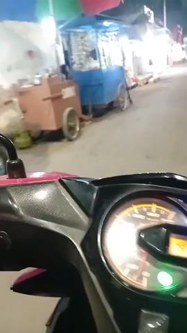 The video shows a first-person perspective of someone riding a motorcycle at night. The motorcycle's dashboard is visible in the foreground, with illuminated gauges. In the background, there are street vendors with stalls and carts, illuminated by lights. People are seen in the background near the stalls. The motorcycle appears to be moving along a paved street.