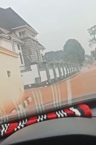 The video shows a view from inside a car, looking out through the windshield towards a large, white and grey two-story house with a dark roof. The house has a fence made of white pillars with grey accents. There are trees in the background and the sky is overcast and hazy. The road appears to be wet. The bottom of the windshield is visible, with a decorative striped material on the dashboard. The car appears to be moving slowly. The car horn is honking.