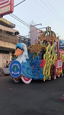 A parade float shaped like a large white swan with a blue and orange beak is moving down a street. The float is decorated with blue and green swirling patterns. Behind the swan is a carriage-like structure with gold accents and a crown on top. Two people are visible riding on the carriage. The float is adorned with colorful flowers and scrollwork. A banner on the side of the float reads "INDONESIA". The background shows buildings and overhead power lines. A person is filming the float with a smartphone.