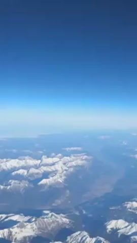 The video shows a view from a high altitude, possibly from an airplane, overlooking a vast landscape of snow-capped mountains. The sky is a clear, vibrant blue at the top of the frame, fading slightly towards the horizon due to atmospheric haze. The mountains are spread out extensively, with varying shades of white snow covering their peaks and ridges. The lower areas between the mountains appear to have patches of green and brown, suggesting valleys or lower-altitude terrain. At least one lake can be seen, nestled within the mountains. The video is stable, without any noticeable camera movement, providing a clear and static view of the mountainous landscape.