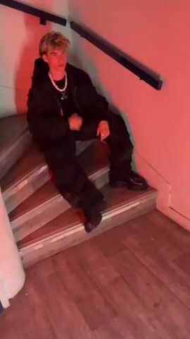 A young man is sitting on wooden stairs in an indoor space with a red light. He is wearing a black jacket, black pants, and black shoes. He has blonde hair and is wearing a silver necklace. The walls are white, and there is a black railing on the wall next to the stairs. The floor is made of wood.