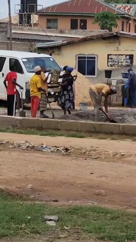 A group of five adults are working at a construction site. Two men are on the left, one wearing a red shirt and black pants, the other in a yellow shirt and red pants. Both are holding shovels. A woman in a black and white dress carries a bucket on her head. A person in a pink top stands near the van. A man in a yellow shirt and brown pants is mixing concrete with a shovel. There is a white van parked on the left. The ground is dirt and there are piles of sand and gravel. The background shows buildings and a water tank.
