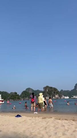 The video shows a beach scene on a sunny day with a clear blue sky. In the foreground, there is a sandy beach with a few items of clothing or towels on the sand. The middle ground features a body of water with numerous people swimming or wading. Several red buoys are visible in the water, marking a boundary. In the background, there are lush green, rocky islands rising from the sea, characteristic of a karst landscape. Several white boats, including what appear to be tour boats or cruise ships, are anchored or moving in the water near the islands. The people in the water are of various ages and genders, some are standing, others are partially submerged, and some are actively swimming. On the beach, there are a few individuals walking along the shore. One person in the foreground is wearing a conical hat and a bikini, carrying a bag. Another person is walking towards the water. The overall atmosphere appears to be that of a popular tourist destination and a recreational beach.