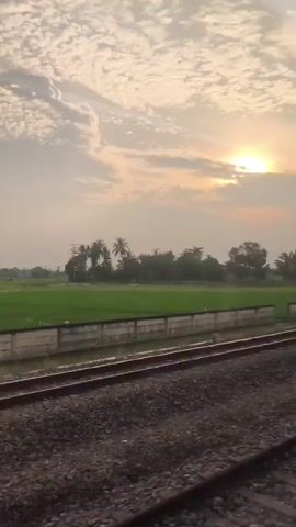 The video shows a view from a train window. Outside the window, there are train tracks in the foreground, with a green field and trees in the background. The sun is setting or rising, casting a warm glow over the scene. The sky is filled with clouds. There are no people or animals visible in the video.