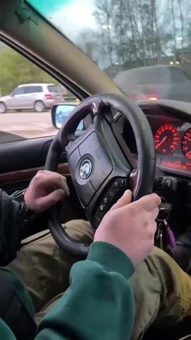The video shows the interior of a BMW car with the driver's perspective. The driver, wearing green pants and a green sweatshirt, is behind the wheel with his hands moving as if he is steering the car. The dashboard includes an aftermarket touchscreen display and red lights on the speedometer and other gauges. A rearview mirror is visible with a hanging air freshener. Outside the car window, other vehicles and buildings are seen. The video seems to be taken during a car ride in an urban area.