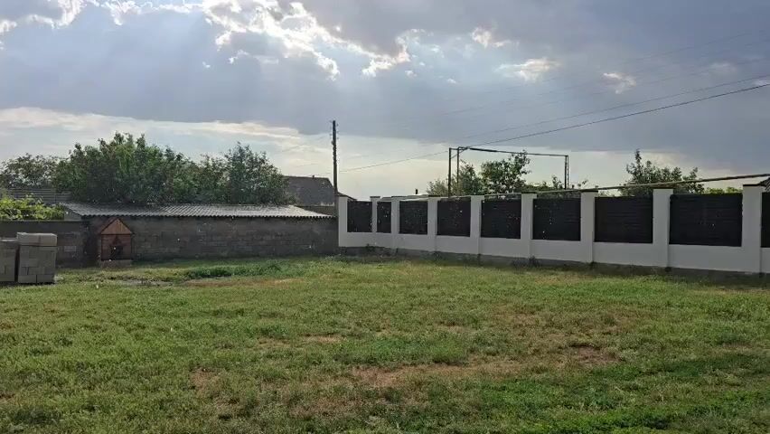 This media file shows an outdoor scene on a cloudy day in Butor, Moldova. The camera is positioned in a grassy yard, looking towards a long, white fence with dark panels. Beyond the fence, there are trees and some buildings, including a smaller structure with a dark roof and a small, enclosed area that might be a doghouse. A stack of grey cinder blocks is visible in the foreground to the left. The sky is filled with dramatic, sunlit clouds, suggesting either the beginning or end of a storm. The sun is breaking through the clouds, casting rays of light downwards. The overall atmosphere is somewhat serene, despite the cloudy and potentially stormy weather. There is no visible text in the frame.