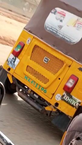The camera is inside a yellow three-wheeled vehicle, possibly a tuk-tuk or auto-rickshaw, driving along a paved road. Outside the vehicle, the landscape features a substantial amount of red-orange dirt or soil. Concrete barriers and building debris are scattered along the roadside. Other vehicles, including a maroon minivan and a white sedan, are visible driving in the opposite direction. The weather appears overcast and bright. The vehicle is moving forward, and the view shifts slightly as it progresses along the road.