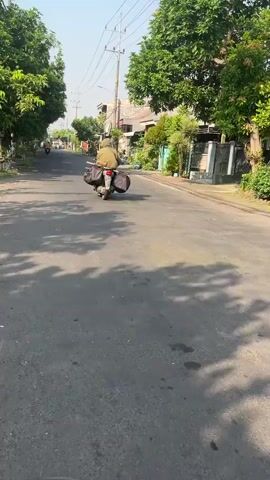 The video shows a person riding a motorcycle down a paved road. The motorcycle is loaded with two large saddlebags on either side of the rear wheel, suggesting it is equipped for travel or carrying cargo. The rider is wearing a helmet and a jacket, obscuring their identity. The road appears to be in a semi-urban or rural setting, with houses and trees visible along the sides. The sun is shining, casting shadows on the road, indicating it is daytime. The overall atmosphere is one of movement and travel.