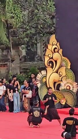 A group of people are gathered outdoors, watching a performance on a red carpet stage. Two individuals are actively participating in the performance, dressed in traditional dark clothing with ornate gold accents. One performer is crouched low, while the other stands with arms outstretched, both appearing focused and engaged.

Behind them, a large, elaborate golden and green decorative structure resembling stylized flames or waves stands against a dark backdrop. Spectators are visible in the background, some standing and others seated, observing the event. The setting appears to be a public space, possibly a cultural festival or a traditional ceremony, given the attire and the decorative elements. The weather seems to be overcast or it's late afternoon, as there are no harsh shadows. The atmosphere is one of attentive observation.

Some individuals in the background are wearing name tags or lanyards, suggesting an organized event. One person in the foreground on the right wears a black t-shirt with yellow lettering, though the text is not clearly legible. Another performer in the background also wears a black t-shirt with similar yellow lettering and a bag slung over their shoulder. The overall impression is of a vibrant cultural presentation in Denpasar, Indonesia.
