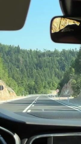 The video captures a first-person perspective of driving on a paved road through a mountainous, forested landscape. The road is a two-lane highway with white lane markings and a guardrail on the right side. The scene is dominated by a dense forest of evergreen trees covering the steep slopes of the mountains. The sky is clear and blue, suggesting a sunny day.

On the right side of the road, there are a couple of road signs. One is a circular sign with a red border and a white background, depicting two cars side-by-side, indicating a prohibition on overtaking. Another sign further down the road is a diamond-shaped warning sign.

The time of day appears to be daytime, likely late morning or early afternoon, given the bright sunlight and clear sky. The weather seems fair and warm, suitable for driving and outdoor activities. The location is identified as Donja Brezna, Montenegro, a region known for its natural beauty and mountainous terrain.

The overall mood conveyed by the video is one of peaceful travel through a scenic natural environment. There are no people directly visible in the frame, and the focus is on the road and the surrounding landscape. The car's dashboard is partially visible at the bottom of the frame, indicating the perspective of a driver. The reflection in the rearview mirror shows a rocky, sunlit hillside, further emphasizing the mountainous setting.