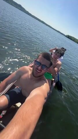 The video shows four young men in a canoe on a lake. The man closest to the camera is shirtless, wearing sunglasses and dark shorts, and taking a selfie video. Behind him, another shirtless man wearing green shorts is paddling. Further back, two other men are visible, one wearing a white shirt. The water is choppy, and the sun is shining. Trees line the shore in the distance. The men appear to be enjoying themselves and paddling the canoe across the lake.