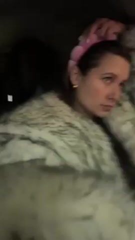 A woman with long brown hair wearing a pink headband and a gray and white faux fur coat sits in the passenger seat of a car. She looks at the camera, adjusts her headband, and makes a kissing face. The car appears to be driving at night.
