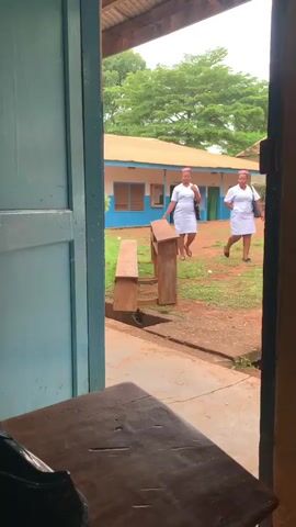 The video shows a view through an open doorway of a building. Outside, there is a courtyard with red soil and grass. Two young women in white dresses and pink headscarves are standing in the courtyard. A low wooden structure, possibly a bench, is in the foreground. In the background, there is a single-story building with blue trim around the windows and doors. Trees with green foliage are visible beyond the building. The lighting suggests it is daytime, possibly overcast.