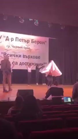 The video shows a performance on a stage in an auditorium, with a banner in Bulgarian visible in the background reading "SU 'Dr. Petar Beron' gr. Cherven Bryag 'All peaks are achievable!'". Five young girls are on stage, all wearing dark pants and light-colored tops. Initially, their arms are raised, but one girl then unfurls a white fabric, holding it out wide. An adult male is standing to the left of the girls on stage, watching them. Several people are seated in the red auditorium seats, with a laptop visible on one of the seats. The lighting is predominantly pink.