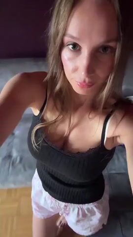 A woman is recording a selfie video in what appears to be a living room. She is wearing a black tank top with lace trim and pink shorts. The room has wooden flooring and a gray couch. The woman is looking directly at the camera and appears to be speaking or singing.