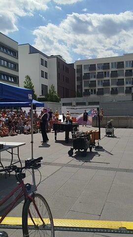 This is a video of a performance taking place outdoors in Offenbach am Main, Germany. The scene is a public square with modern buildings in the background. The sky is blue with fluffy white clouds, suggesting it is a pleasant, sunny day. A crowd of people is gathered, seated on the ground and standing, watching a performance.

The central focus of the performance appears to be a man in a black suit who is conducting or leading the event. In front of him, there is a large, colorful inflatable structure that resembles a giant bubble. There are also several musical instruments set up, including what looks like a piano and drums. Other individuals are visible interacting with the instruments and the inflatable structure.

To the right of the frame, a bicycle is parked, with its front wheel and part of its frame visible. The ground is paved with grey tiles, and there is a yellow line marking a boundary or pathway. A blue tent is also visible in the lower left corner, possibly providing shade or housing some equipment. The overall atmosphere appears to be festive and engaging, with the audience eagerly observing the show. The time of day is likely daytime, given the bright sunlight and shadows. There is no visible text in the media file.