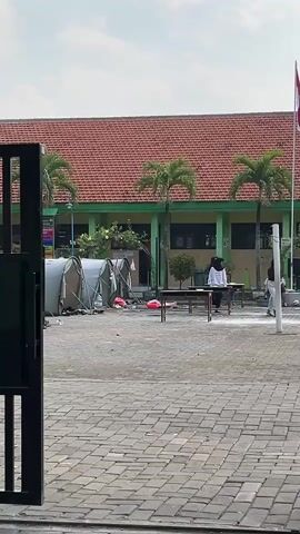 The video shows an outdoor scene in front of a school building with a red tile roof and green trim. In the foreground, a paved area with interlocking bricks is visible, with a black metal gate on the left. Several tents are set up on the left side of the paved area, with some scattered debris in front of them. In the middle ground, there are tables and chairs, with a few people, appearing to be students, interacting around them. One person in a white shirt and dark pants is at a table, and other individuals in dark clothing, some wearing headscarves, are also present. A white line is marked on the ground in front of the tables. On the right side of the scene, a flagpole with a red and white flag is visible. The environment appears to be outdoors on a bright, possibly sunny day, with palm trees and some greenery in the background. The overall impression is of an outdoor event or activity taking place at a school.