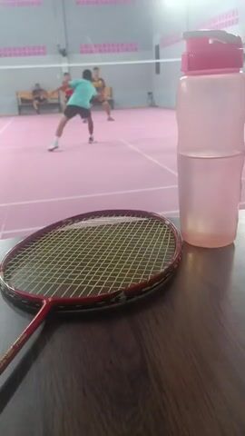 The video shows a badminton court with several people playing. In the foreground, a badminton racket and a water bottle are on a wooden surface. The court is pink, and the net is visible in the background. The people in the background are wearing athletic clothing. The lighting appears to be artificial, typical of an indoor sports facility. The camera is focused on the racket and bottle, with the court and players slightly blurred in the background. The scene suggests a break or downtime during a badminton game.