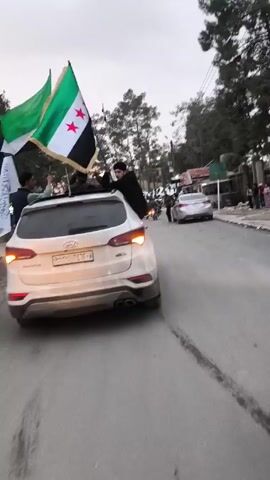 The video shows a street parade in what appears to be a Middle Eastern country. A white SUV is moving slowly along a street. Three people stand on the car’s roof, waving Syrian flags. The vehicle’s license plate is visible. A man is seen precariously leaning out of the sunroof. The street is crowded with motorcycles carrying passengers. A few pedestrians are visible on the sidewalks. Other vehicles, including cars and a white van, are also on the street. The day appears overcast. Buildings and trees line the street.