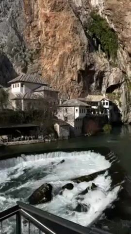 The video shows a scenic view of Blagaj Tekija in Bosnia and Herzegovina.  The camera is positioned on a ledge overlooking a rushing waterfall, with a metal railing visible at the bottom of the frame.  Behind the waterfall is the Tekija, a historic Dervish monastery, built into the base of a tall, rocky cliff face. The monastery buildings are white with stone roofs, and there appear to be people dining at tables outside one of the structures.  The water is turbulent and frothy as it cascades over the man-made steps forming the waterfall.  The rocky cliff is partially covered with some green vegetation.  The sky is not visible, but the scene is brightly lit.